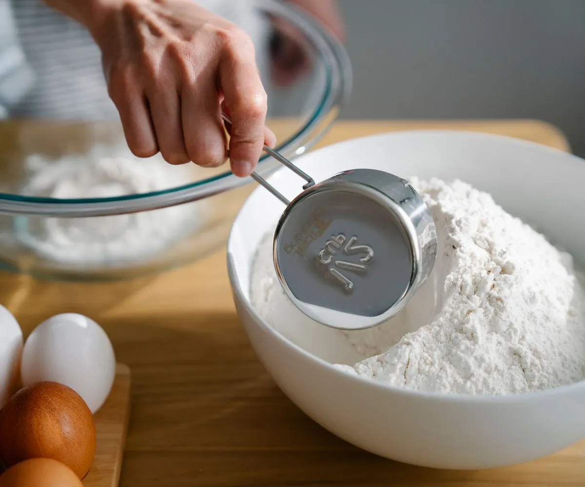 A hand measuring flour into a mixing bowl beside fresh eggs
