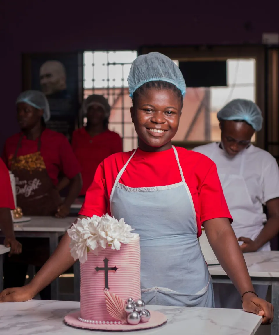 A smiling young baker in an apron and hairnet standing beside a decorated pink cake in a baking class